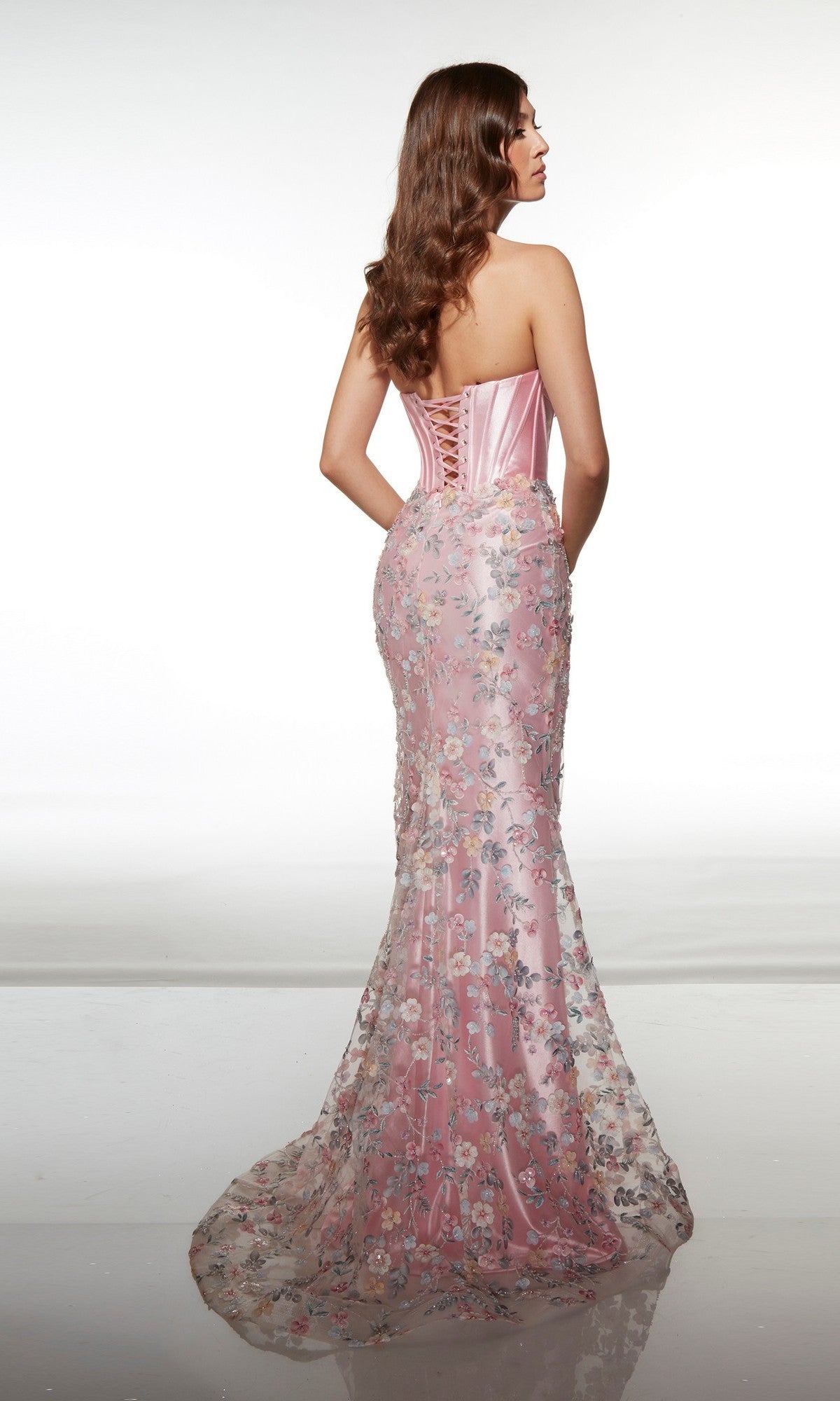 A woman is seen from the back wearing the Alyce 62197 long prom dress, a strapless pink gown with a lace-up corset bodice and a sheer floral overlay adorned with delicate 3D flower embellishments.